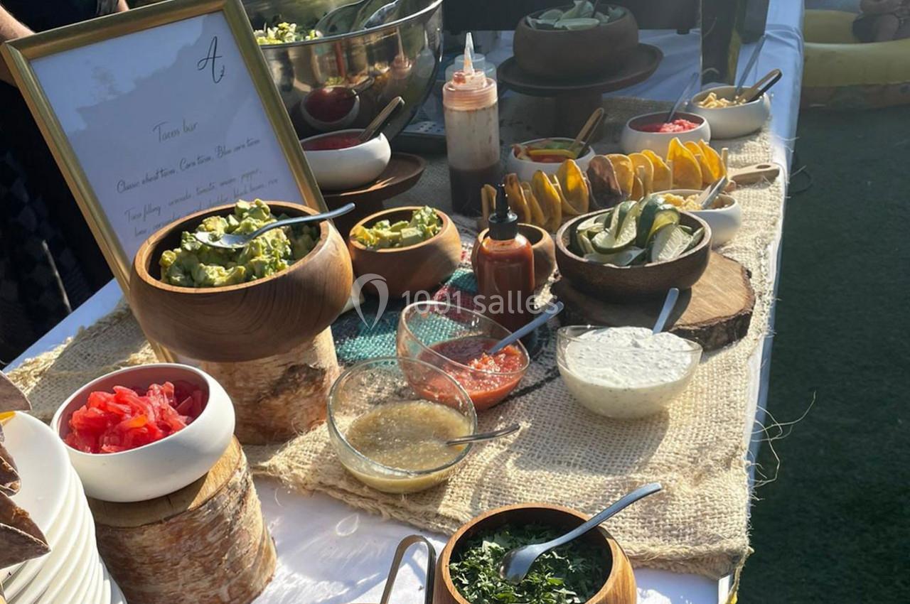 Buffet avec divers condiments, sauces et garnitures disposés sur une table en extérieur, éclairée par le soleil.