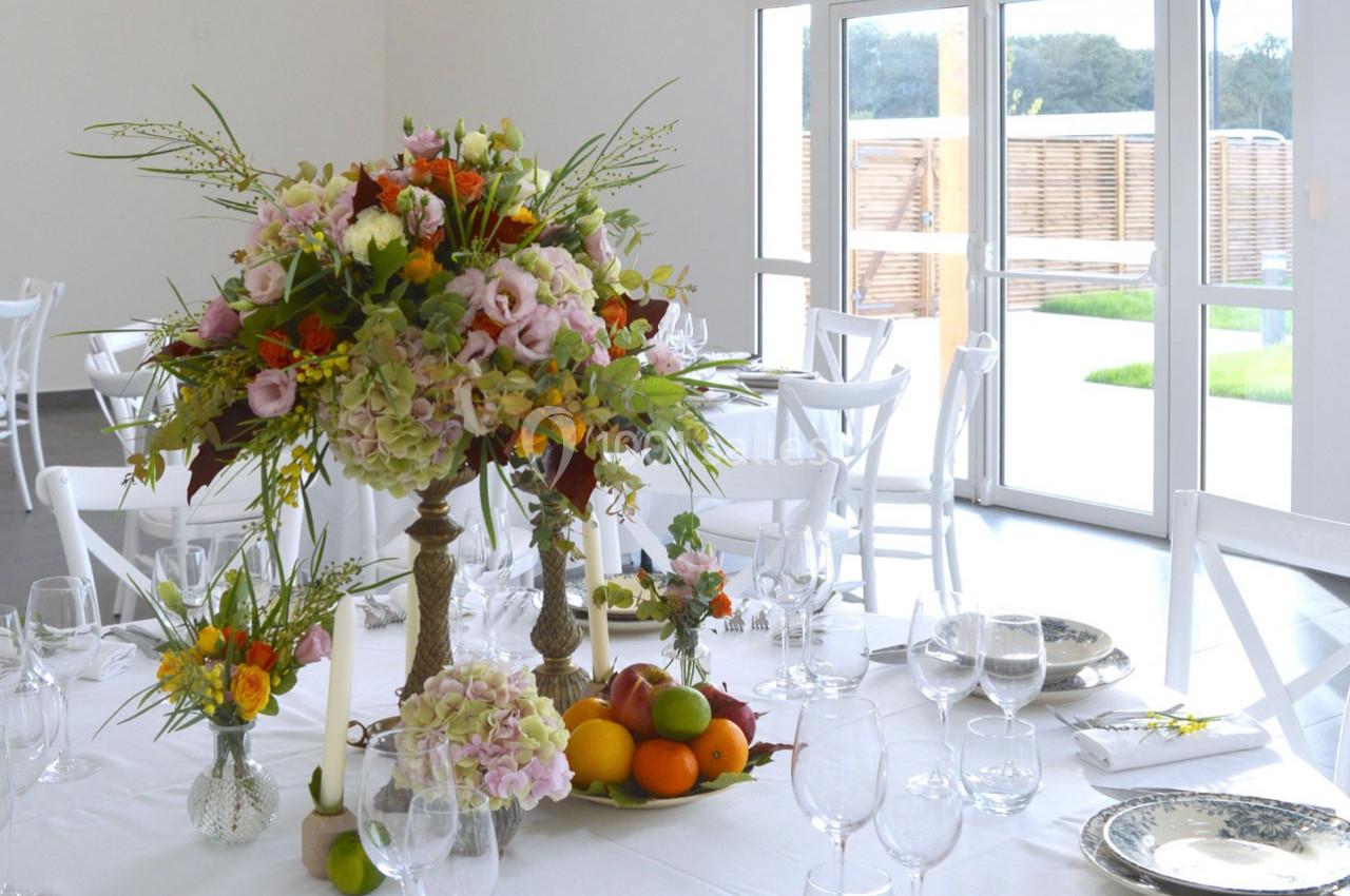 Centre de table floral avec fruits sur une table dressée dans une salle lumineuse aux grandes baies vitrées.