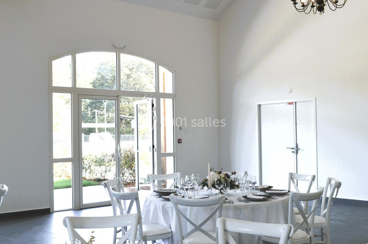 Salle lumineuse avec tables dressées, chaises blanches, poutres apparentes, lustre et grande baie vitrée donnant sur un…