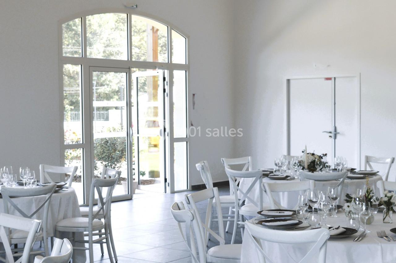 Salle lumineuse avec tables rondes dressées, chaises blanches et grandes fenêtres donnant sur un extérieur verdoyant.