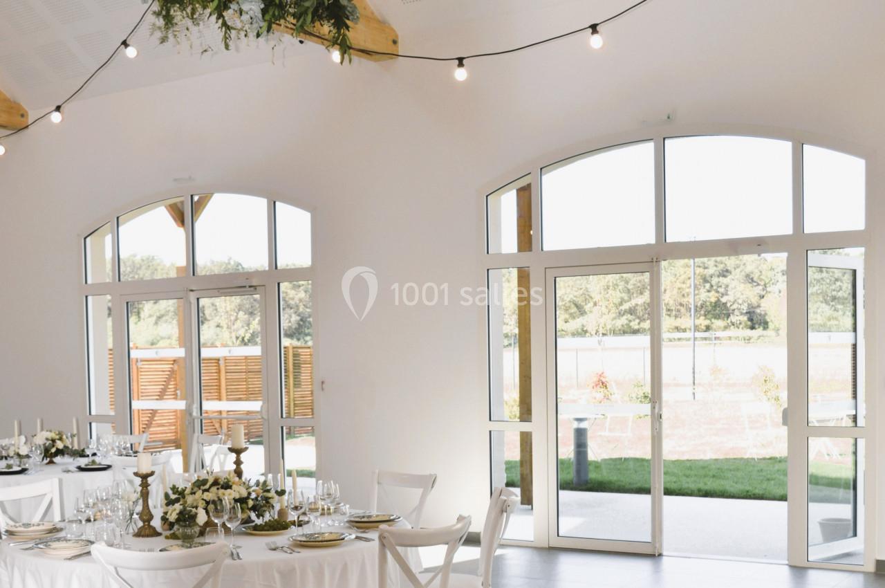 Salle lumineuse avec grandes baies vitrées, tables décorées pour un repas et lustre suspendu au plafond en bois.
