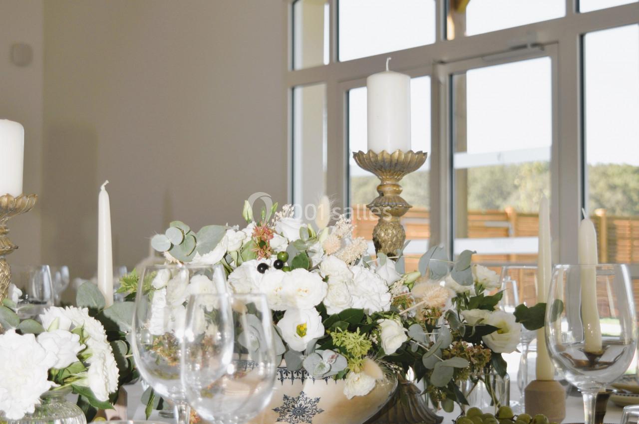 Table élégamment dressée avec des fleurs blanches, bougies et assiettes, dans une salle lumineuse avec baie vitrée.