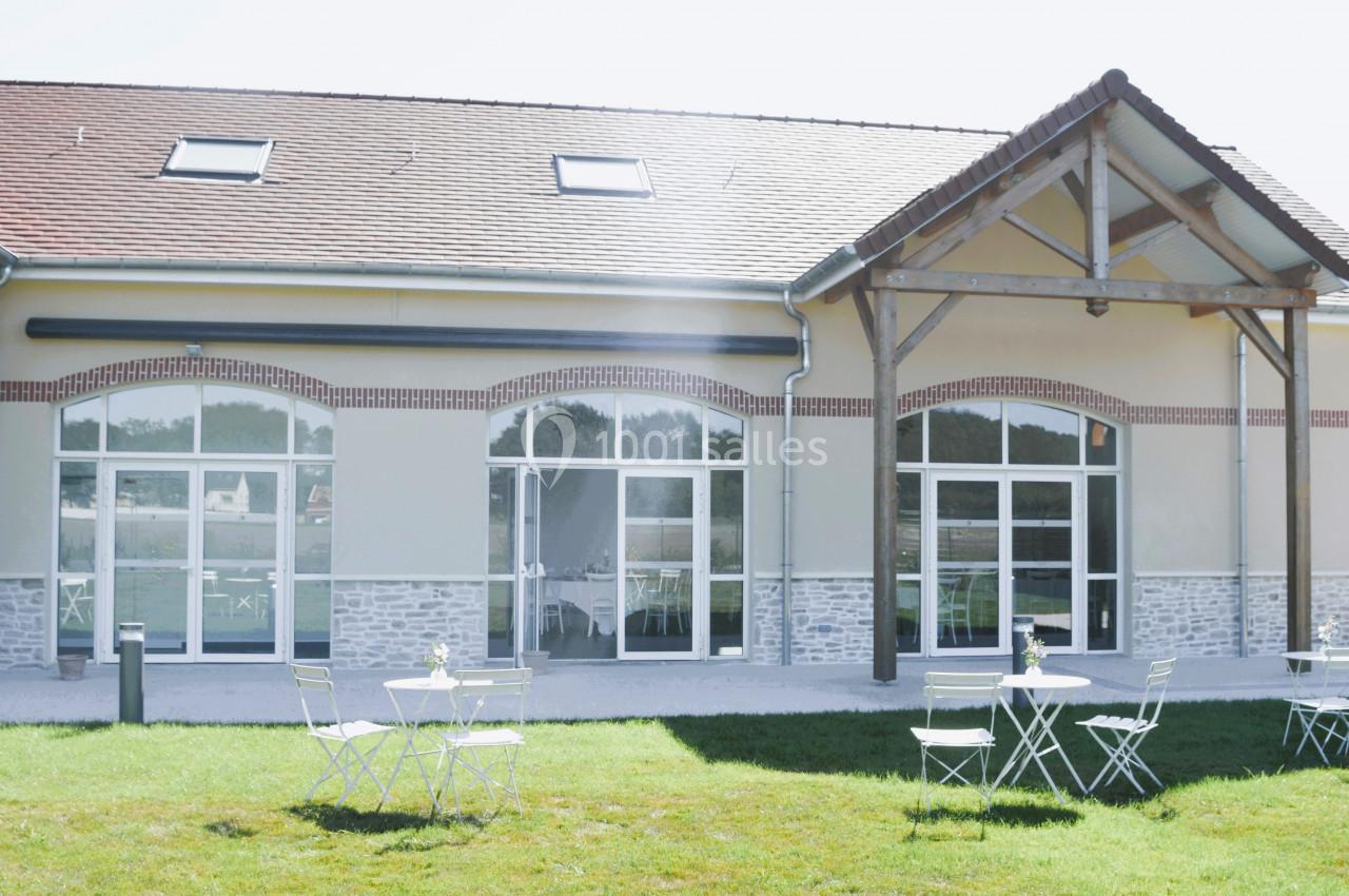 Façade d'un bâtiment moderne avec grandes baies vitrées, terrasse aménagée avec tables et chaises sur pelouse.