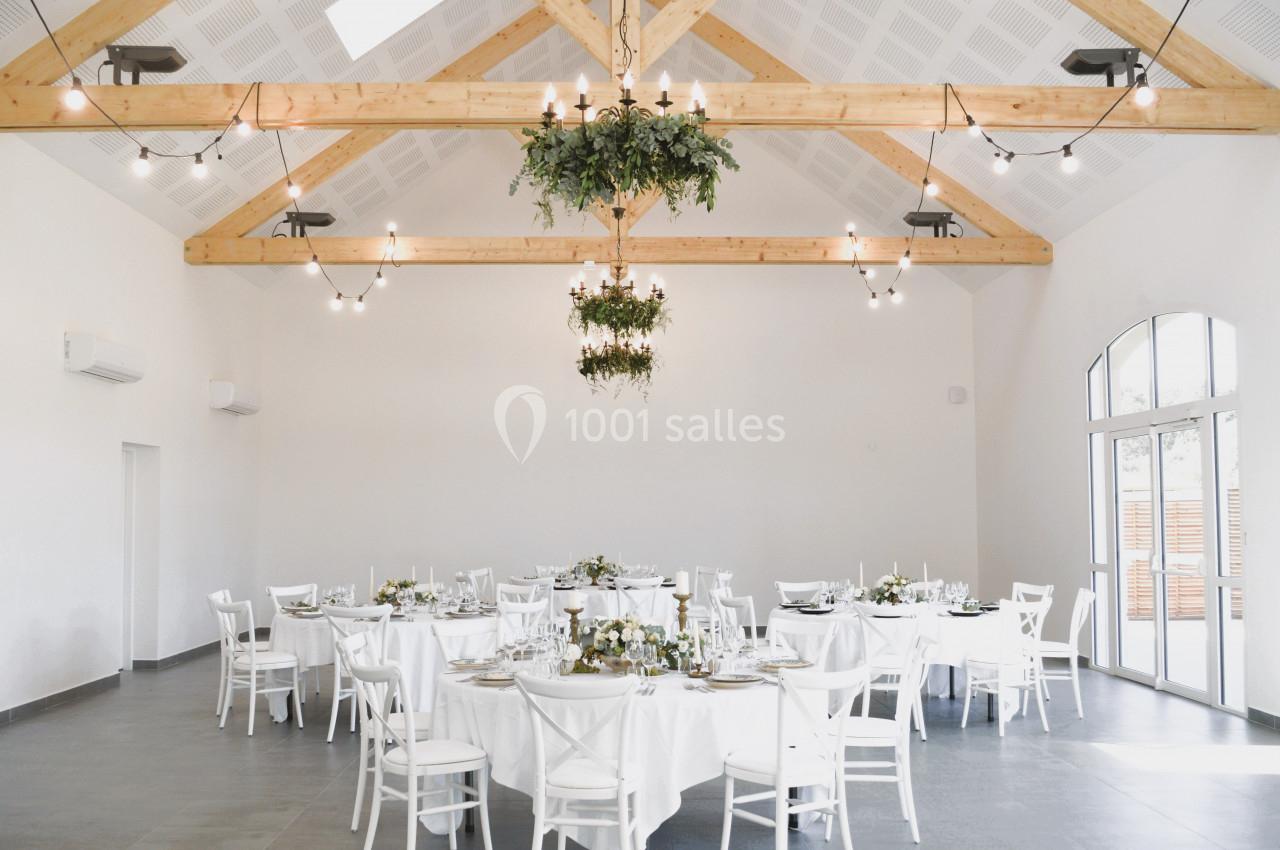 Salle lumineuse avec des tables rondes dressées, des chaises blanches, des poutres apparentes et des suspensions végétales.