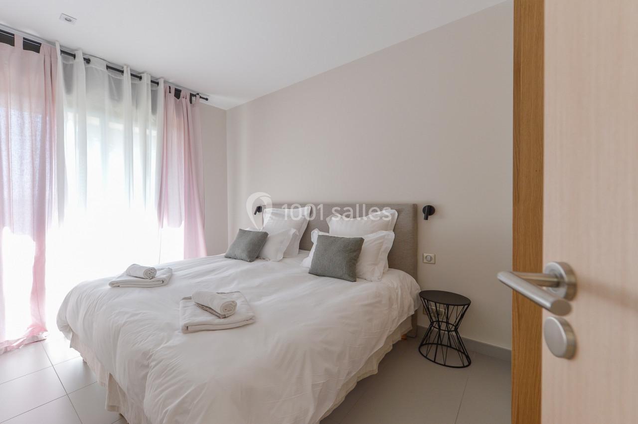 Chambre lumineuse avec un lit double, des draps blancs, des coussins gris, des rideaux clairs et une table d'appoint noire.
