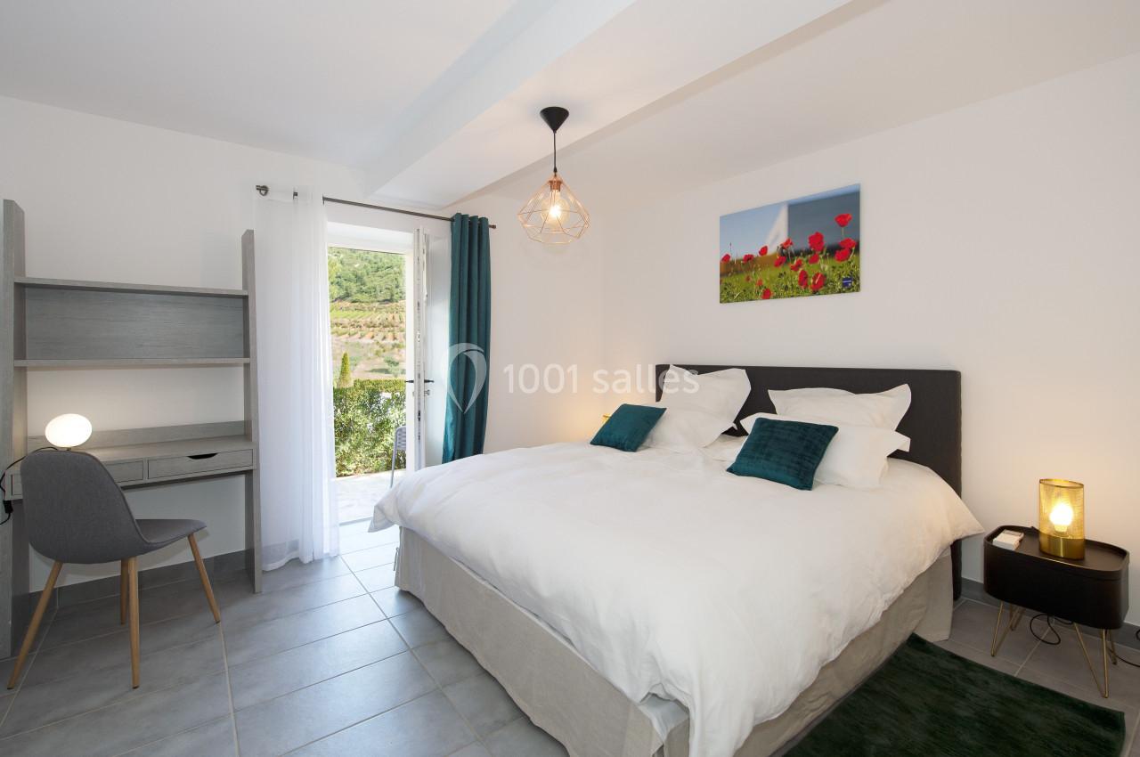 Chambre lumineuse avec un lit double, un bureau, une chaise, une lampe et une porte-fenêtre donnant sur un jardin.