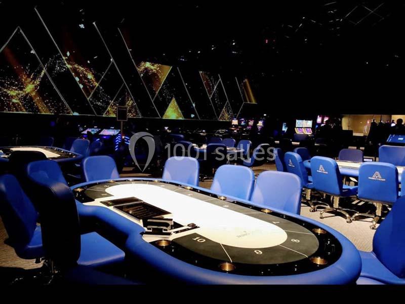 Salle de casino avec tables de jeu, chaises bleues et écrans lumineux en arrière-plan.