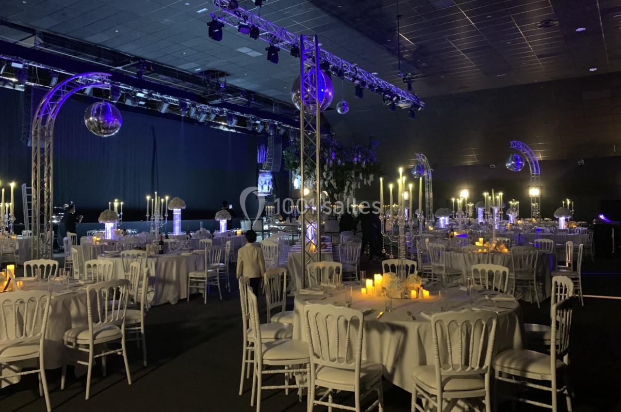 Salle de réception décorée avec des tables rondes, nappes blanches, chandeliers allumés et éclairage tamisé.