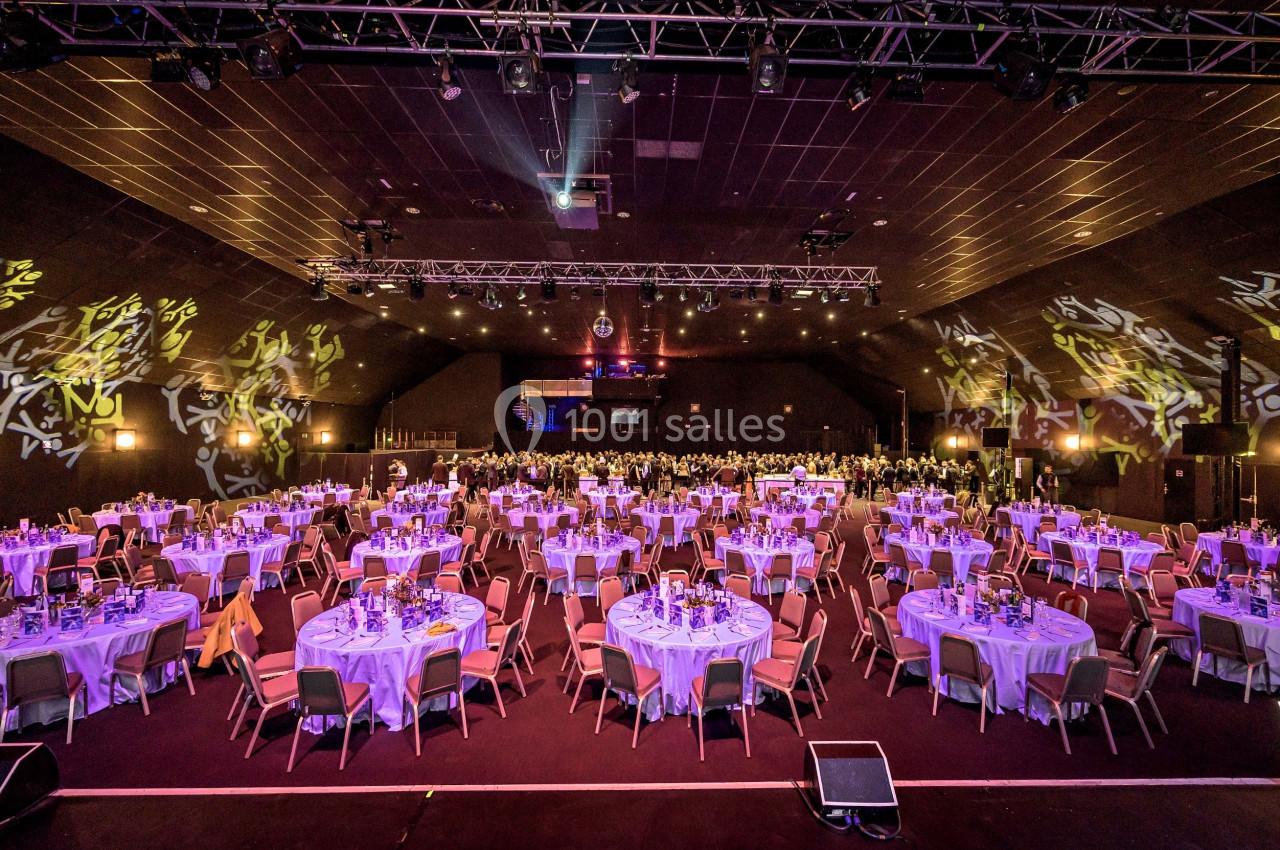 Salle de réception avec de nombreuses tables rondes dressées pour un événement, éclairée par des lumières colorées.