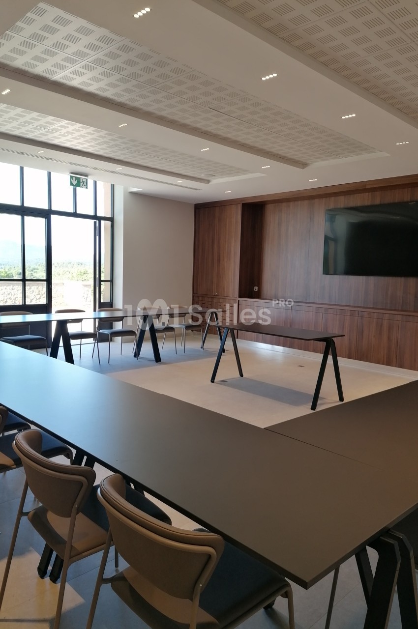 Salle de réunion moderne avec tables en U, chaises, écran mural et grande fenêtre laissant entrer la lumière naturelle.