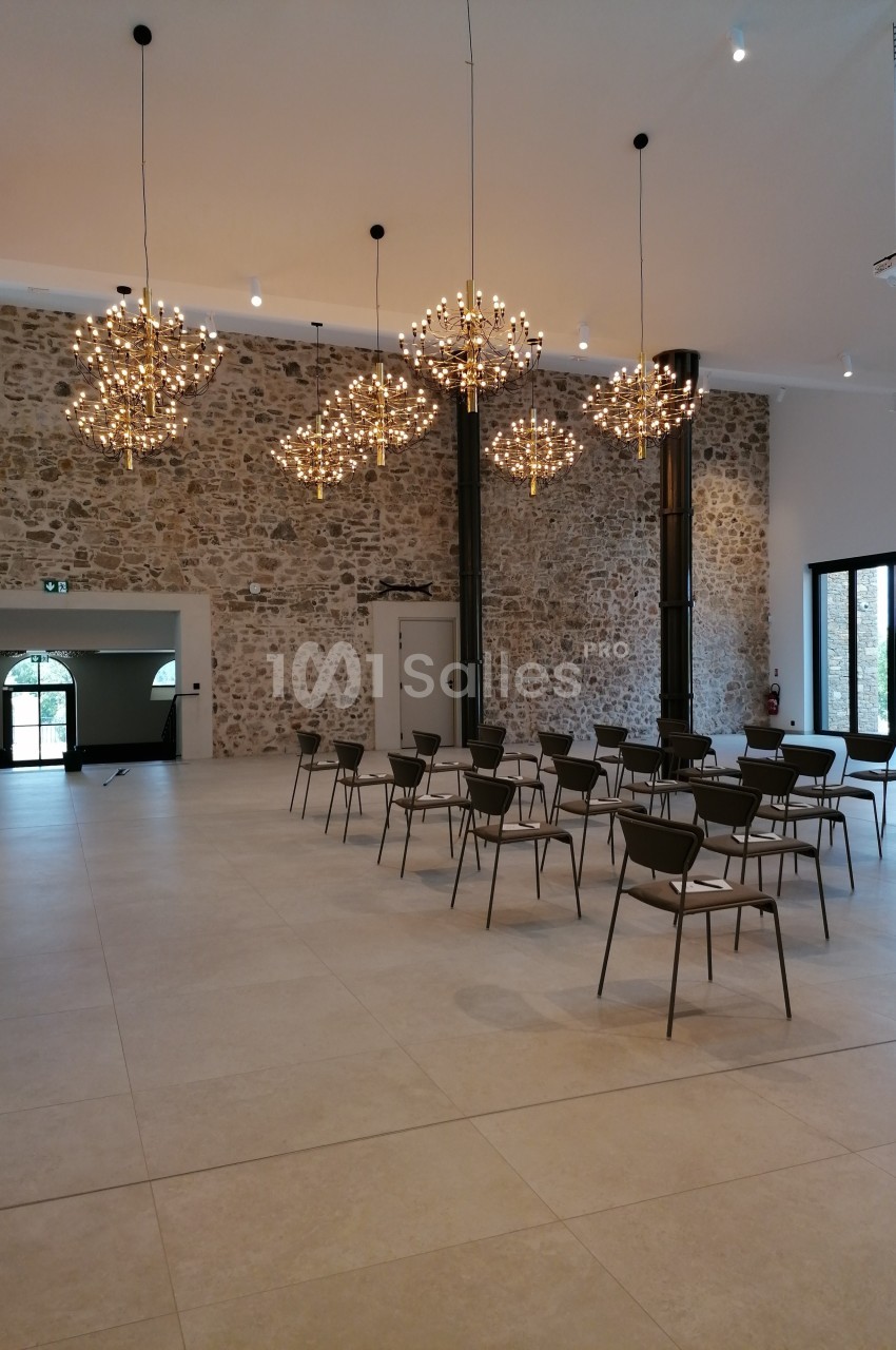 Salle lumineuse avec murs en pierre, chaises disposées en rangées et lustres modernes suspendus au plafond.
