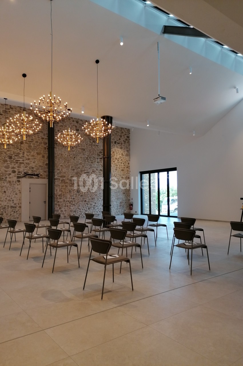 Salle lumineuse avec murs en pierre, chaises disposées en rangées, grandes baies vitrées et lustres modernes suspendus.