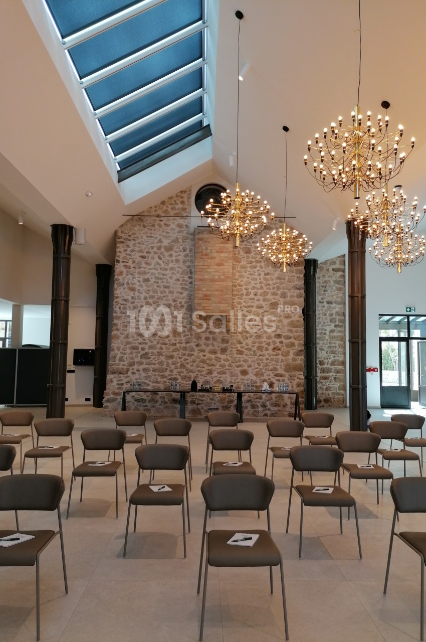 Salle lumineuse avec murs en pierre, chaises disposées en rangées et lustres suspendus sous un plafond vitré.