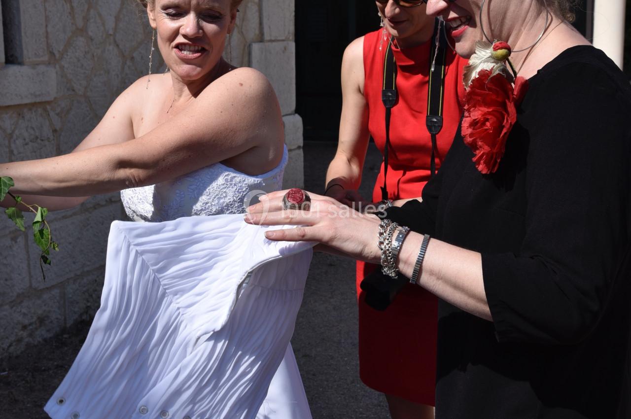 Une mariée ajuste sa robe blanche avec l'aide de deux femmes à l'extérieur d'un bâtiment en pierre.