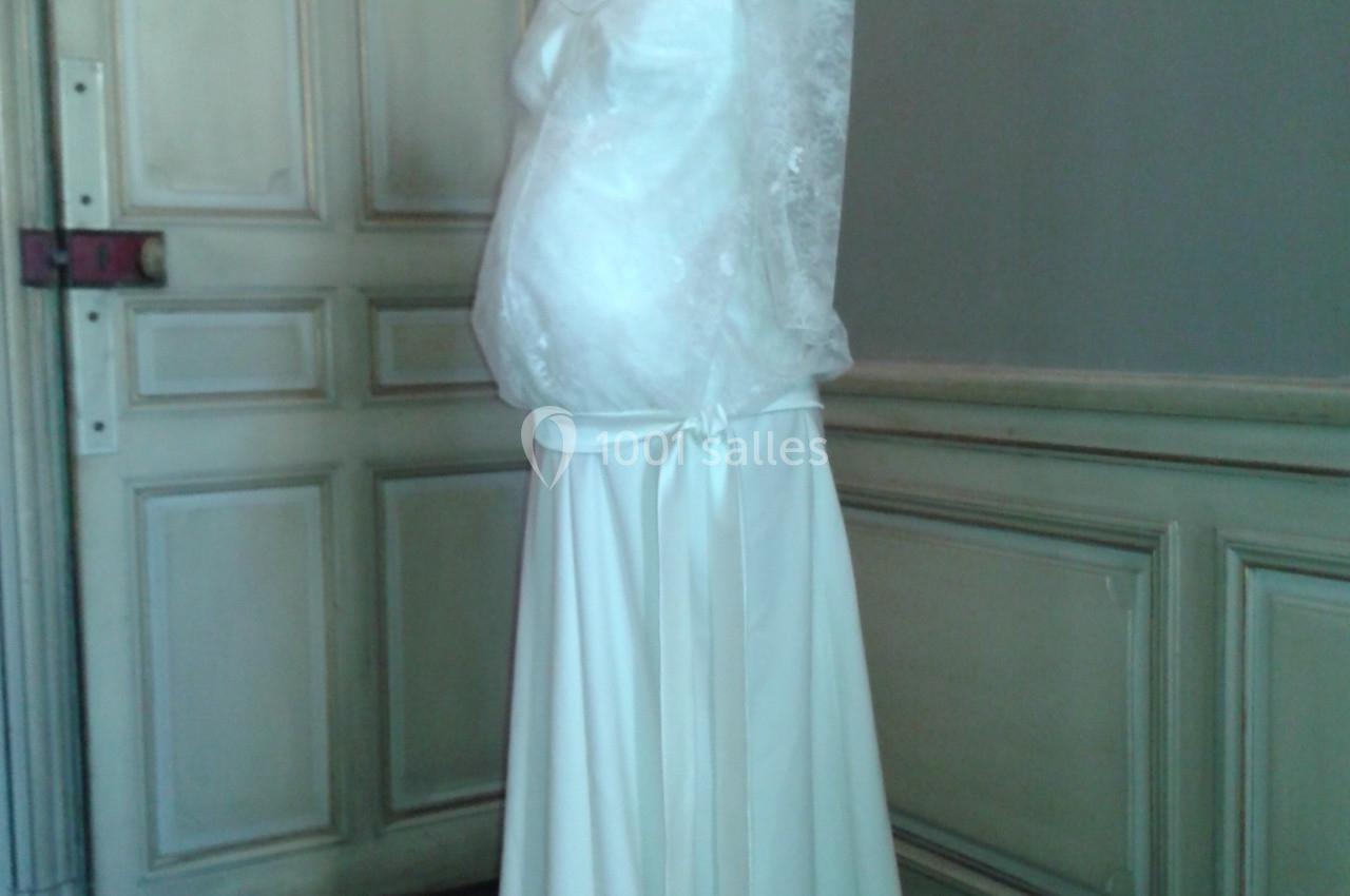 Robe de mariée blanche avec manches longues en dentelle, présentée sur un mannequin dans une pièce au décor classique.