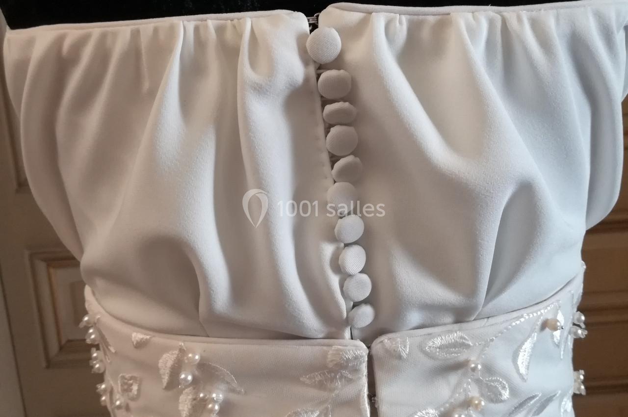 Dos d'une robe blanche avec boutons recouvrant la fermeture et ceinture ornée de broderies florales et perles.