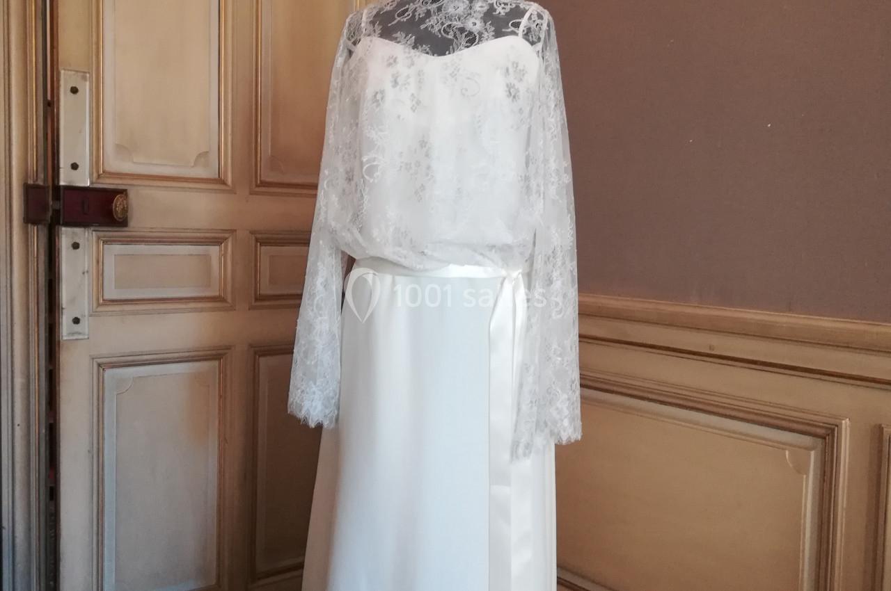 Robe de mariée blanche avec haut en dentelle et manches longues, exposée sur un mannequin dans une pièce ancienne.