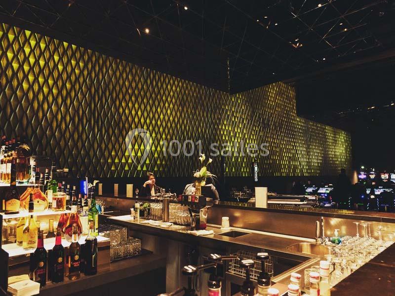 Bar moderne avec comptoir en métal, bouteilles alignées et éclairage mural jaune doré dans une ambiance tamisée.