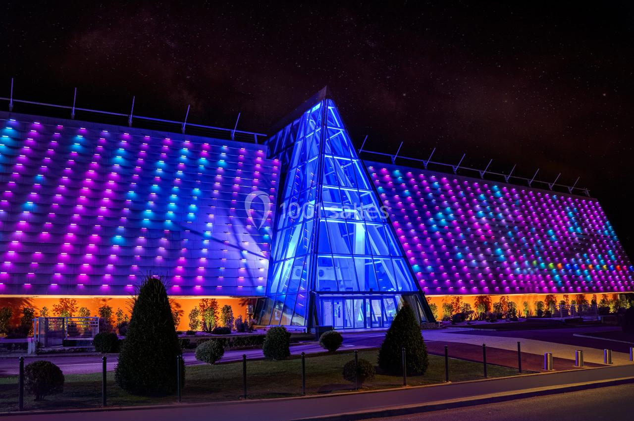 Façade illuminée d'un bâtiment moderne avec des motifs colorés et une entrée vitrée triangulaire, de nuit.