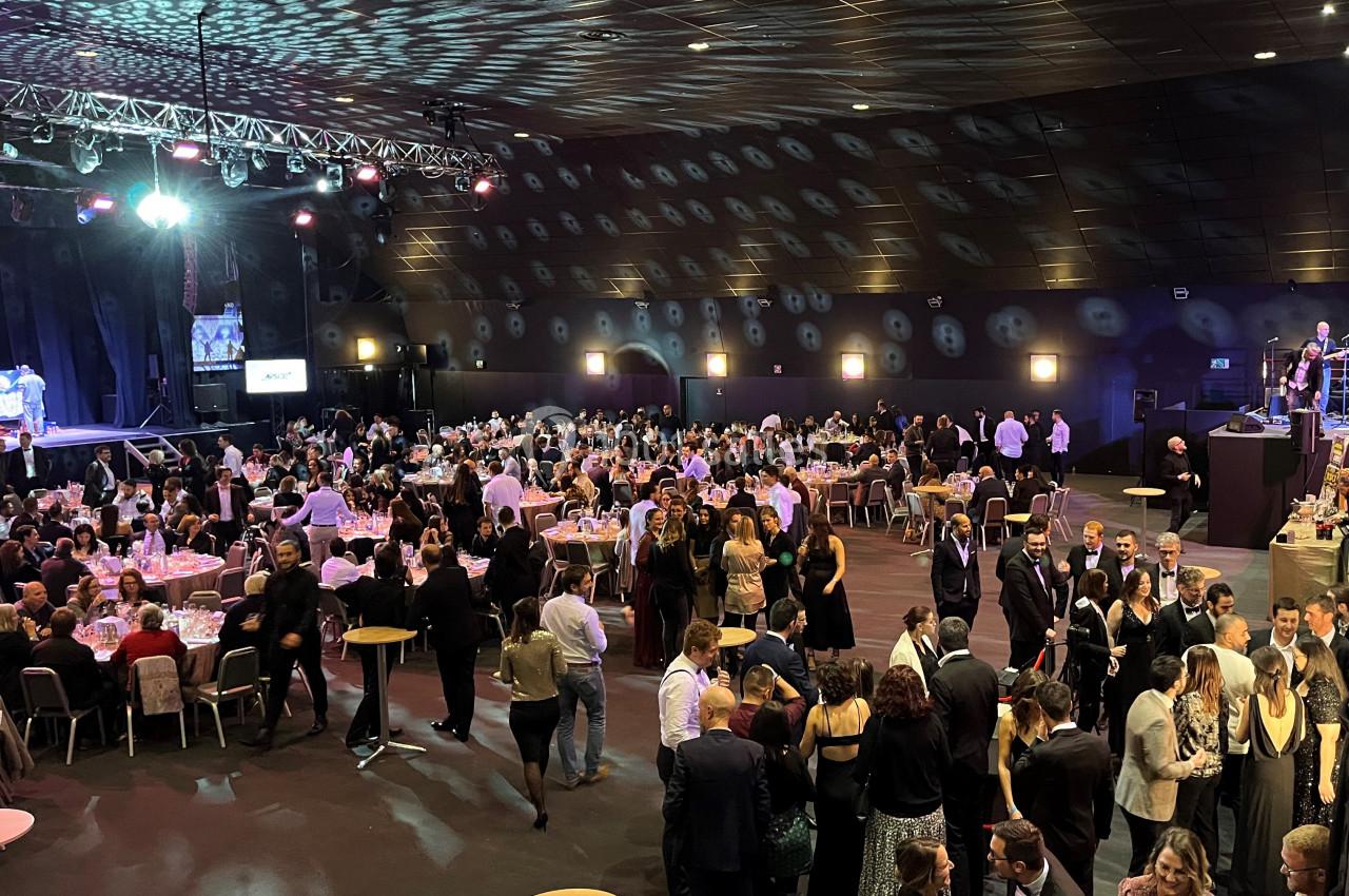 Salle de réception bondée avec des invités en tenue de soirée, assis à des tables ou debout, sous un éclairage tamisé.