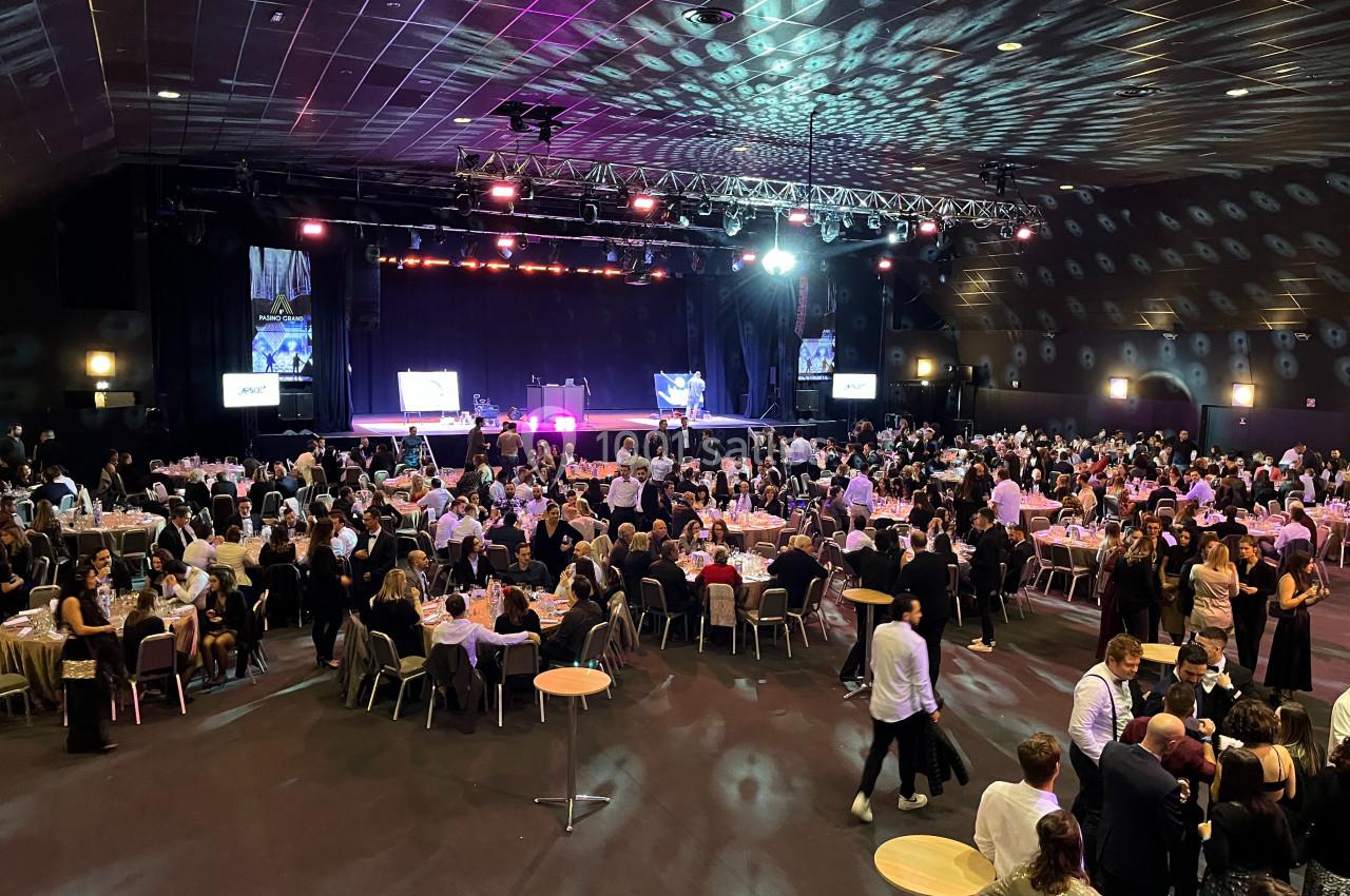 Salle de réception animée avec de nombreuses tables occupées et une scène éclairée pour un événement en cours.
