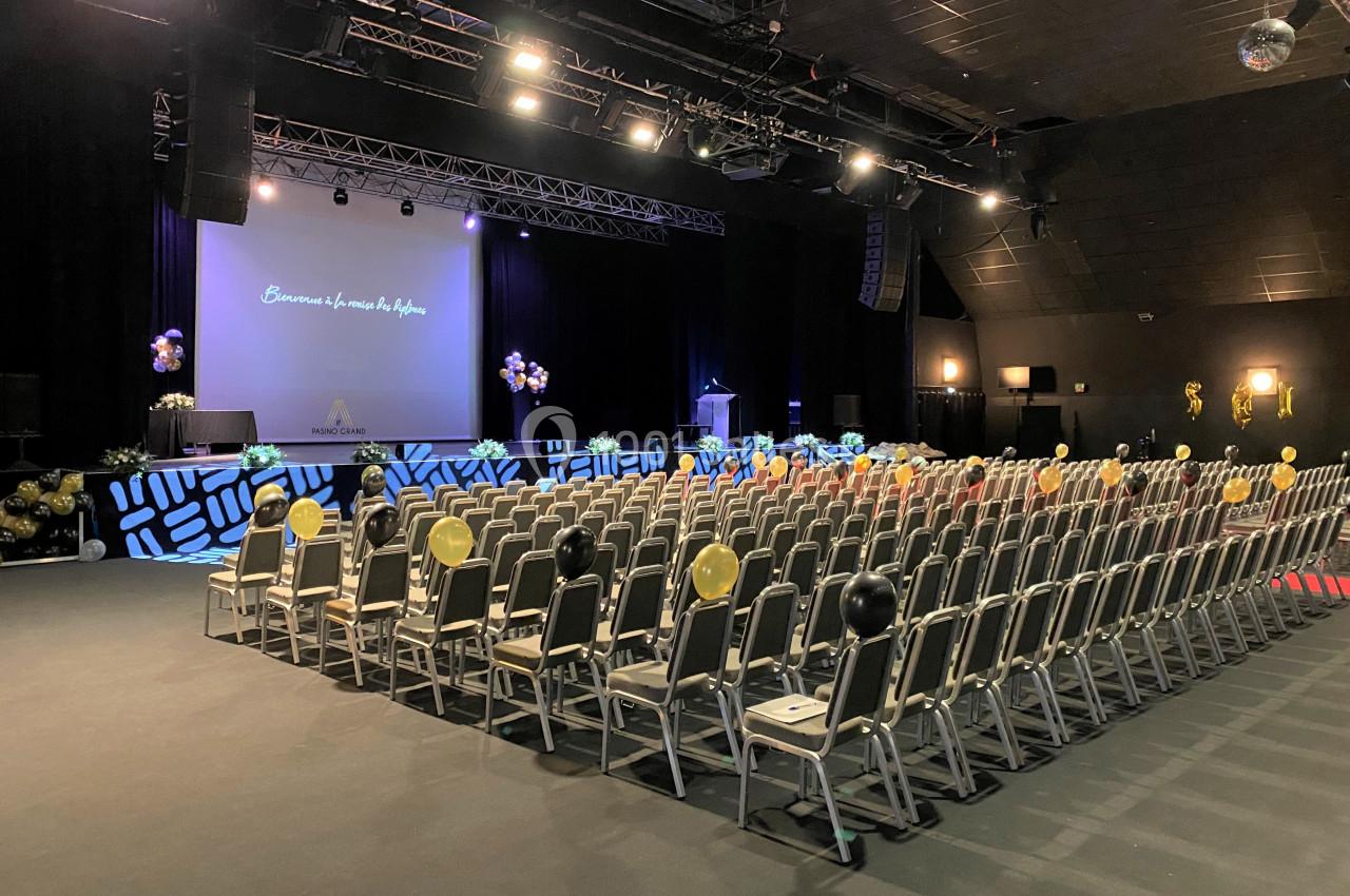 Salle de conférence avec des rangées de chaises, une scène décorée de ballons et un écran de projection allumé.