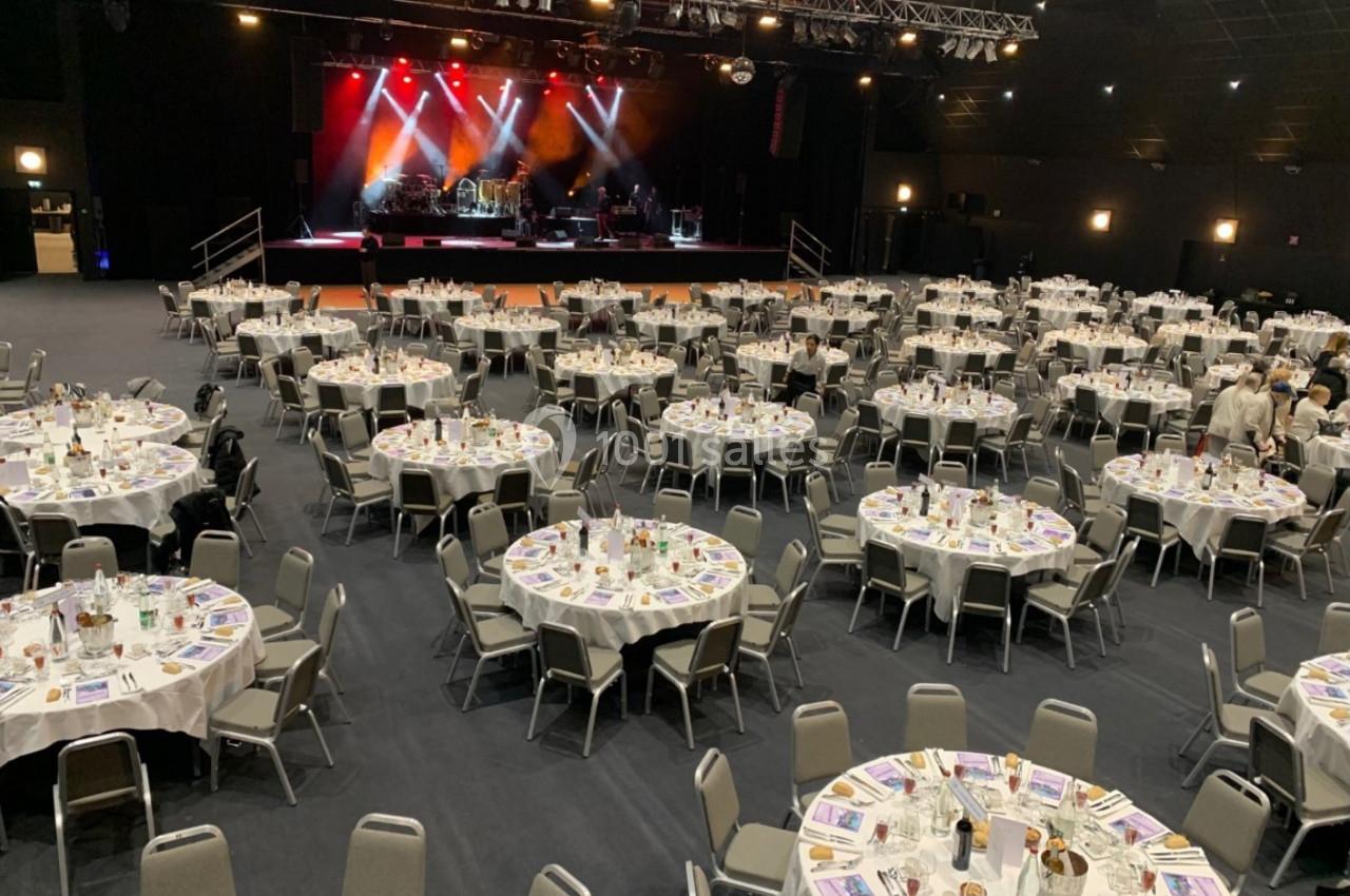 Salle de réception avec des tables rondes dressées pour un événement, scène éclairée en arrière-plan.