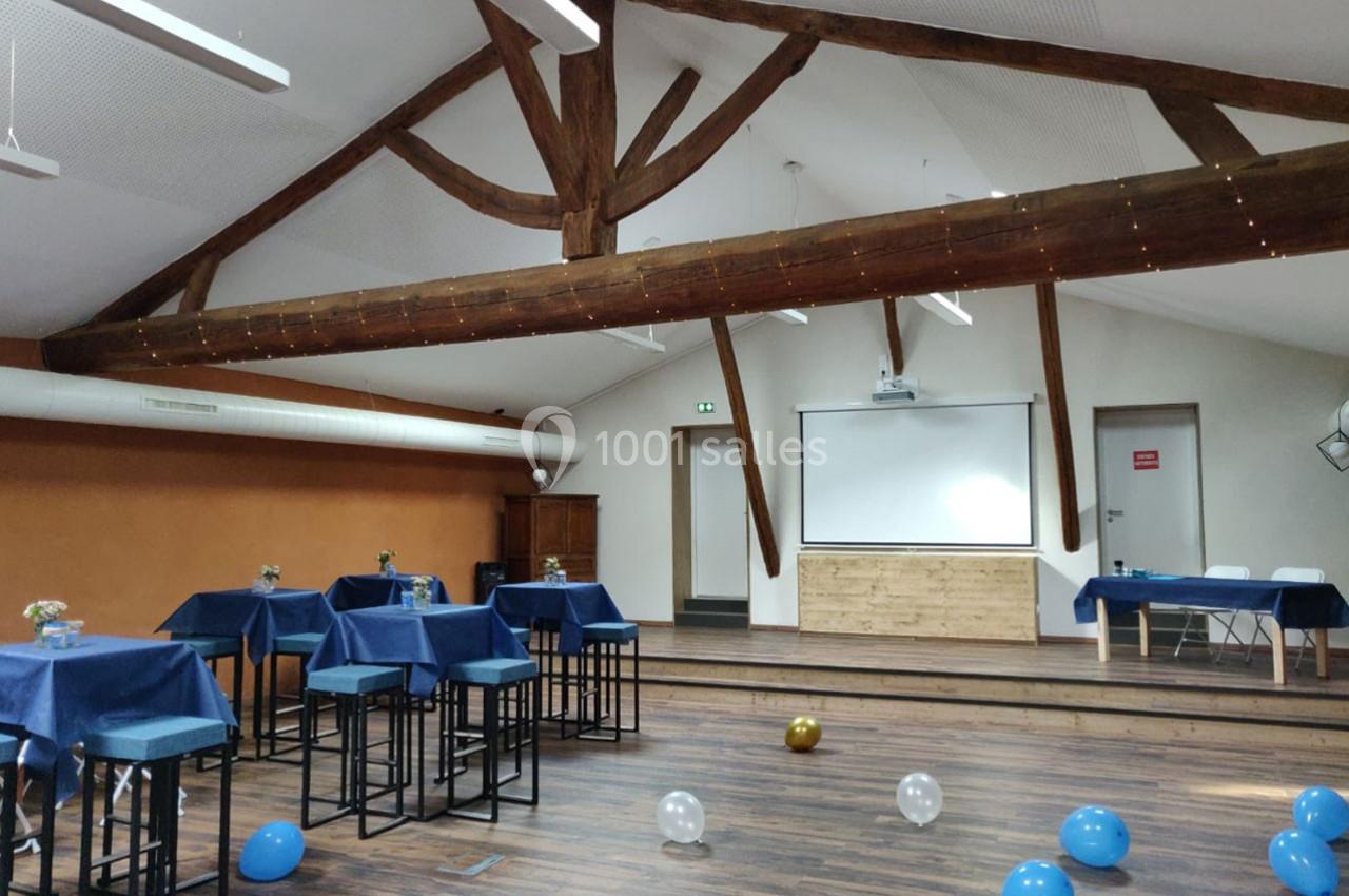 Salle avec poutres apparentes, tables hautes nappées de bleu, écran de projection et ballons au sol.