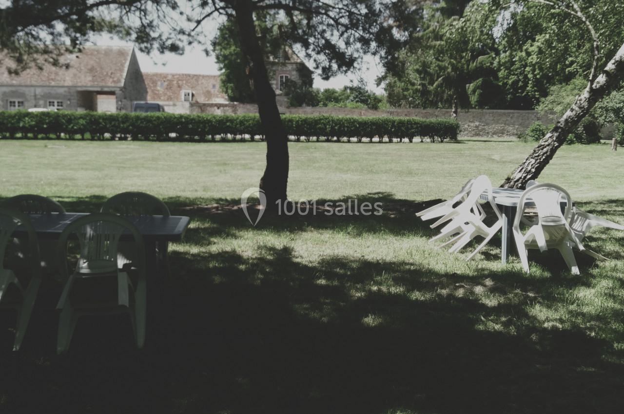 Chaises blanches empilées et tables disposées sur une pelouse ombragée, avec des arbres et des bâtiments en arrière-plan.