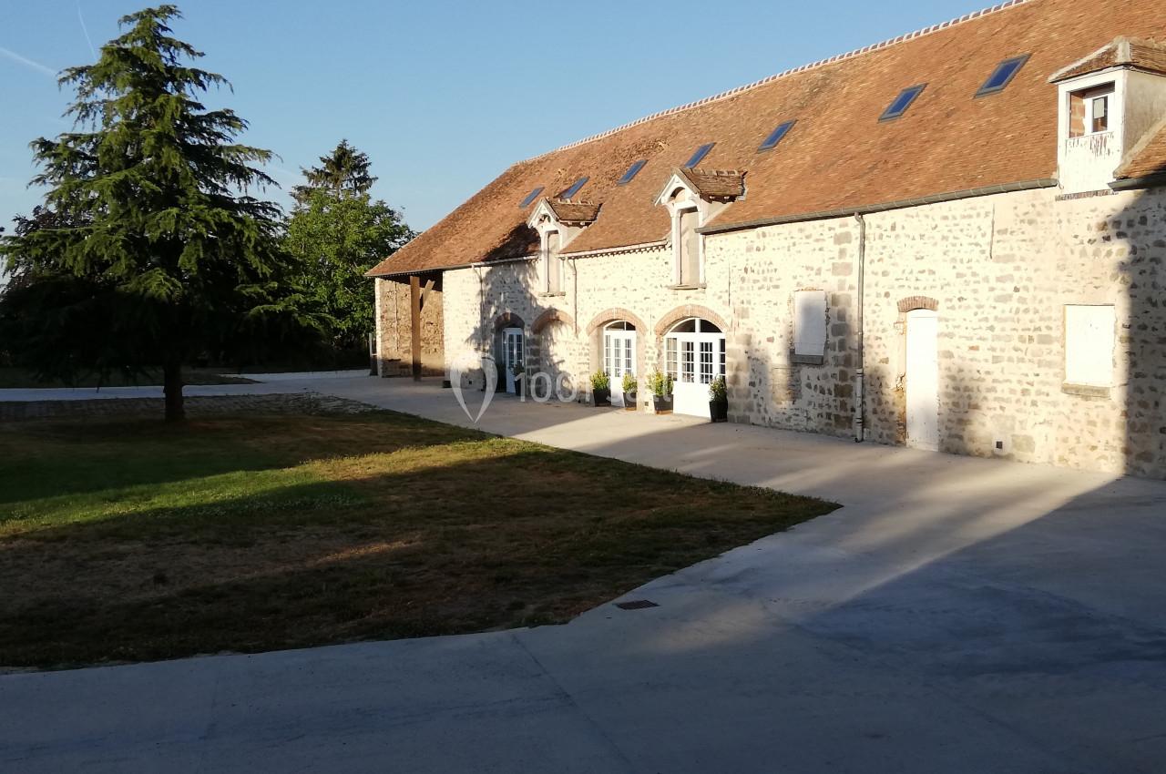 Bâtiment en pierre avec toit en tuiles, fenêtres blanches et cour pavée, entouré de pelouse et d'arbres.