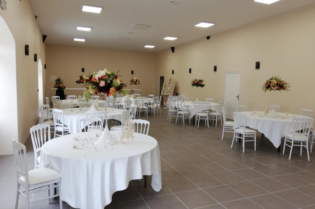 Salle de réception lumineuse avec tables rondes dressées, nappes blanches et décorations florales.
