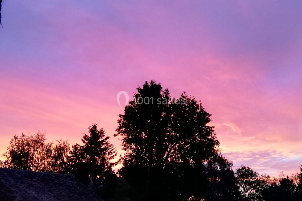 Ciel aux teintes roses et violettes au coucher du soleil, avec des silhouettes d'arbres et de toits au premier plan.