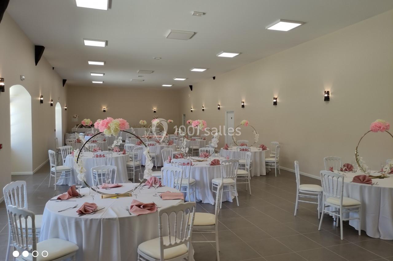 Salle de réception décorée avec des tables rondes, nappes blanches, serviettes roses et centres de table fleuris.