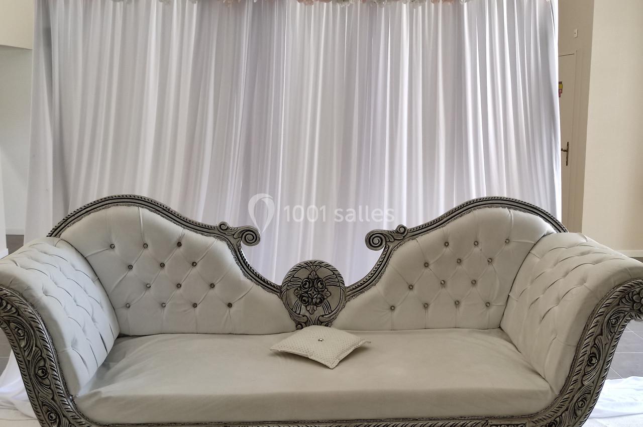 Canapé beige capitonné avec accoudoirs sculptés, placé devant un rideau blanc décoré de fleurs roses et blanches.