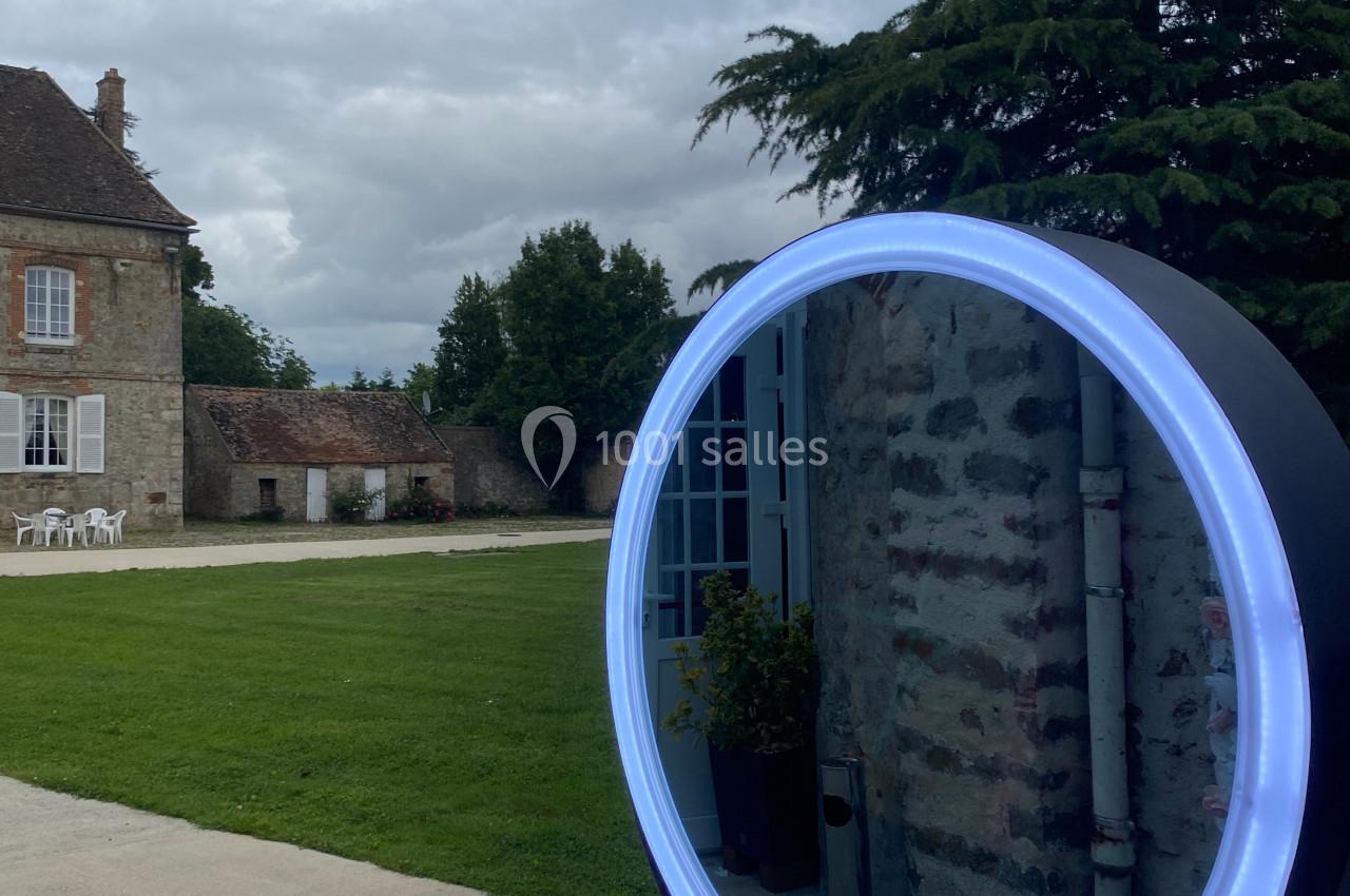 Miroir lumineux circulaire placé à l'extérieur, reflétant un bâtiment en pierre et un jardin sous un ciel nuageux.