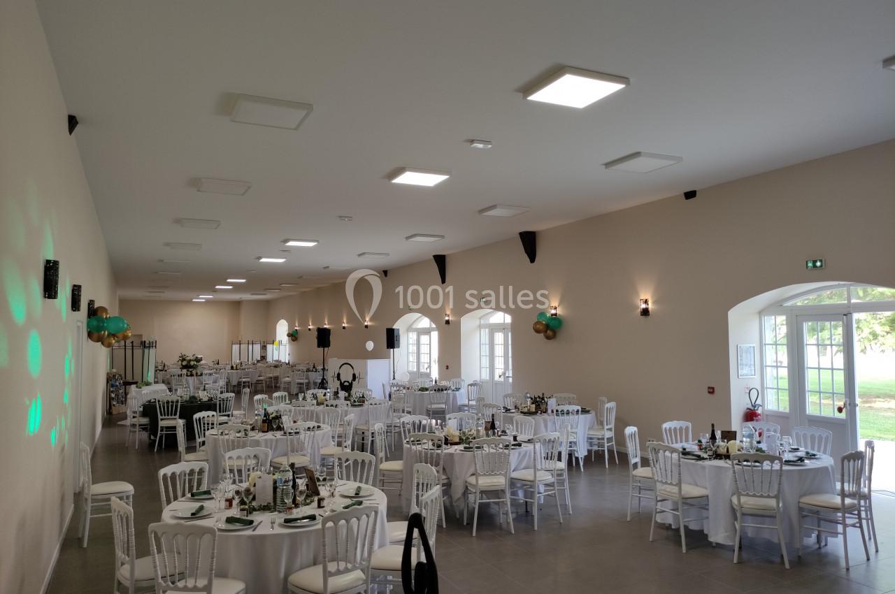 Salle de réception lumineuse avec tables rondes décorées, chaises blanches et ballons accrochés aux murs.