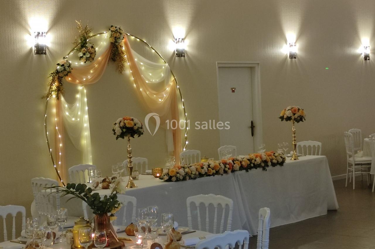 Décoration de salle avec arche lumineuse ornée de fleurs, table d'honneur et chaises blanches disposées autour des tables.