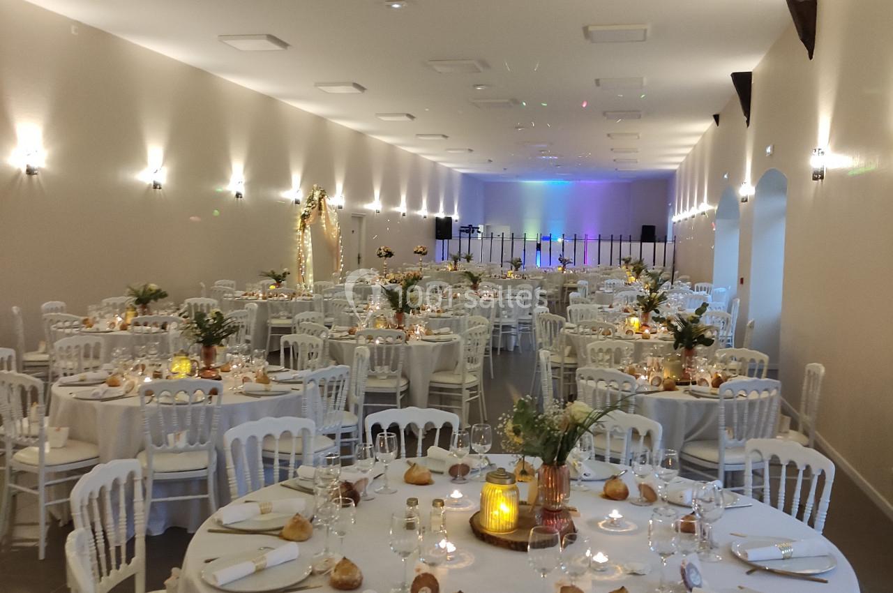 Salle de réception décorée avec des tables rondes dressées, chaises blanches et éclairage chaleureux.