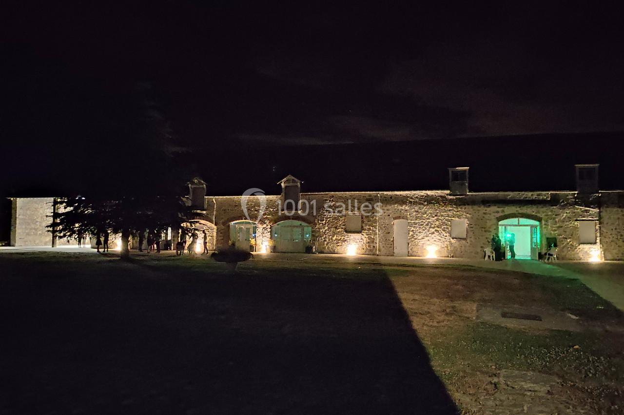 Bâtiment en pierre éclairé la nuit, avec des fenêtres illuminées et une cour partiellement ombragée.