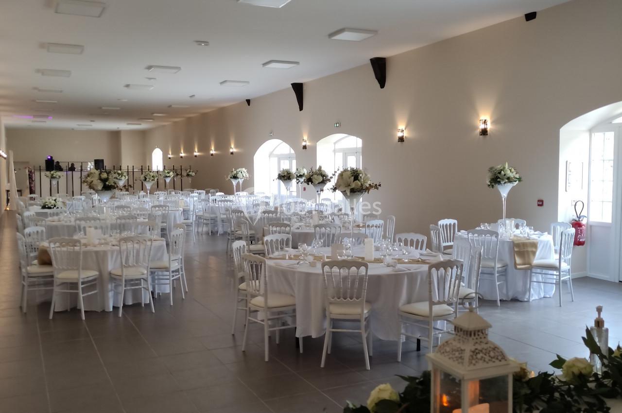 Salle de réception lumineuse avec tables rondes dressées, chaises blanches et décorations florales élégantes.