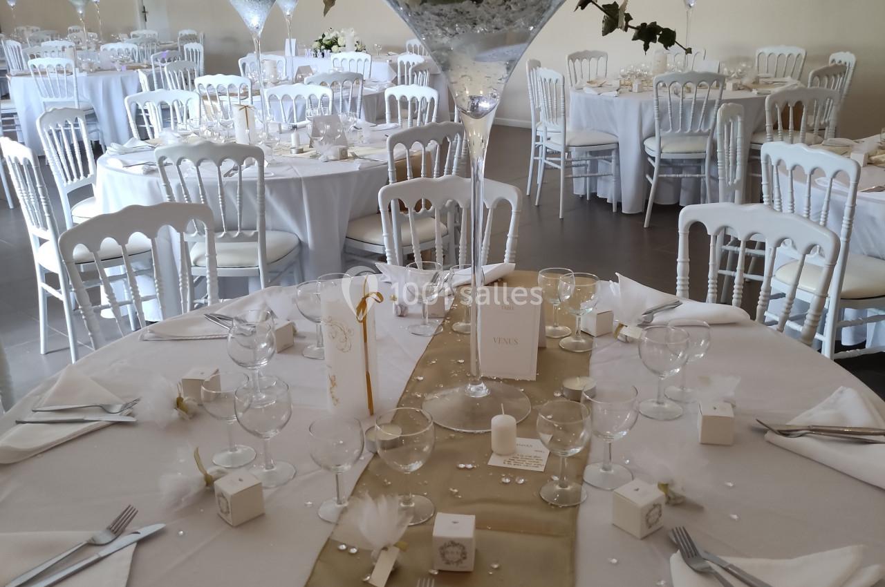 Salle de réception décorée pour un mariage, avec tables rondes dressées, nappes blanches et centres de table fleuris.