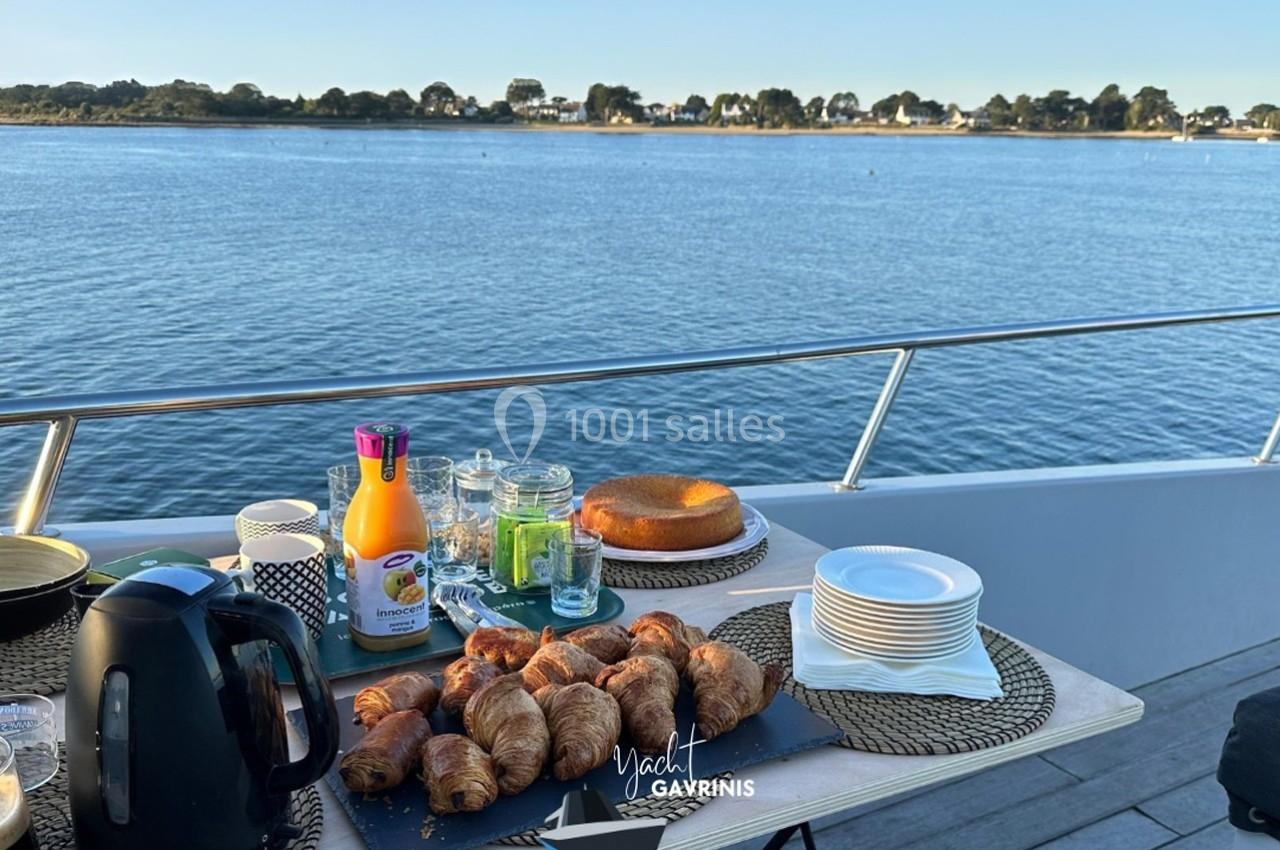 Petit-déjeuner avec croissants, jus d'orange et café servi sur le pont d'un bateau avec vue sur un paysage côtier.