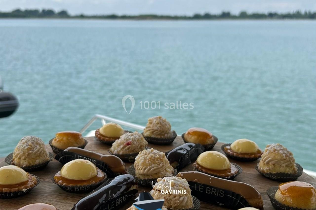 Plateau de pâtisseries variées présenté sur un bateau, avec une vue sur une étendue d'eau et un horizon boisé.