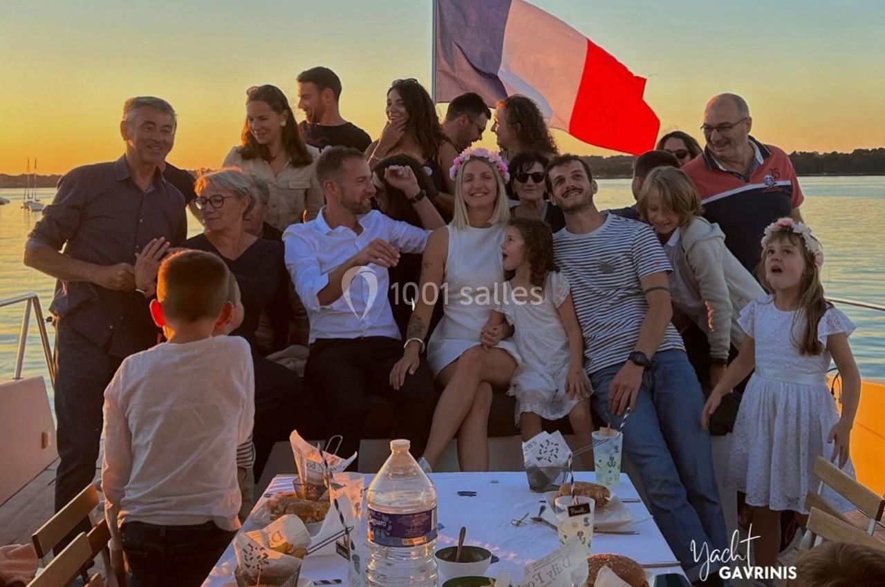 Un groupe de personnes souriantes assises sur un bateau au coucher du soleil, avec un drapeau français en arrière-plan.