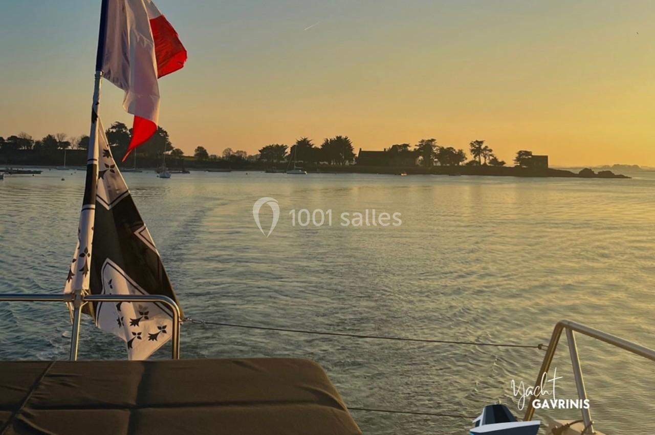 Vue depuis un bateau au coucher du soleil, avec un drapeau français flottant et un rivage arboré à l'horizon.