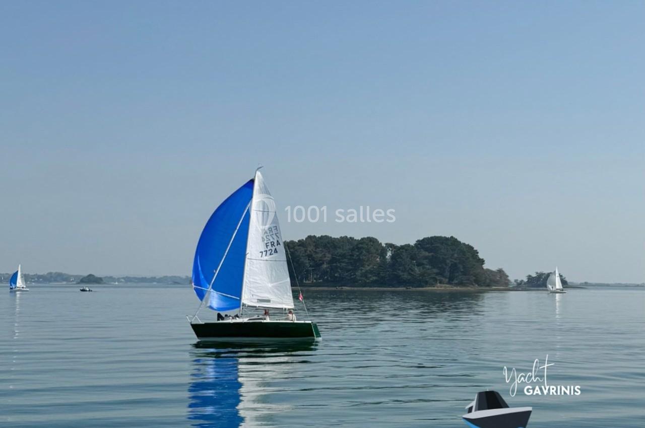 Voilier avec une voile bleue naviguant sur une mer calme, avec une île boisée en arrière-plan.