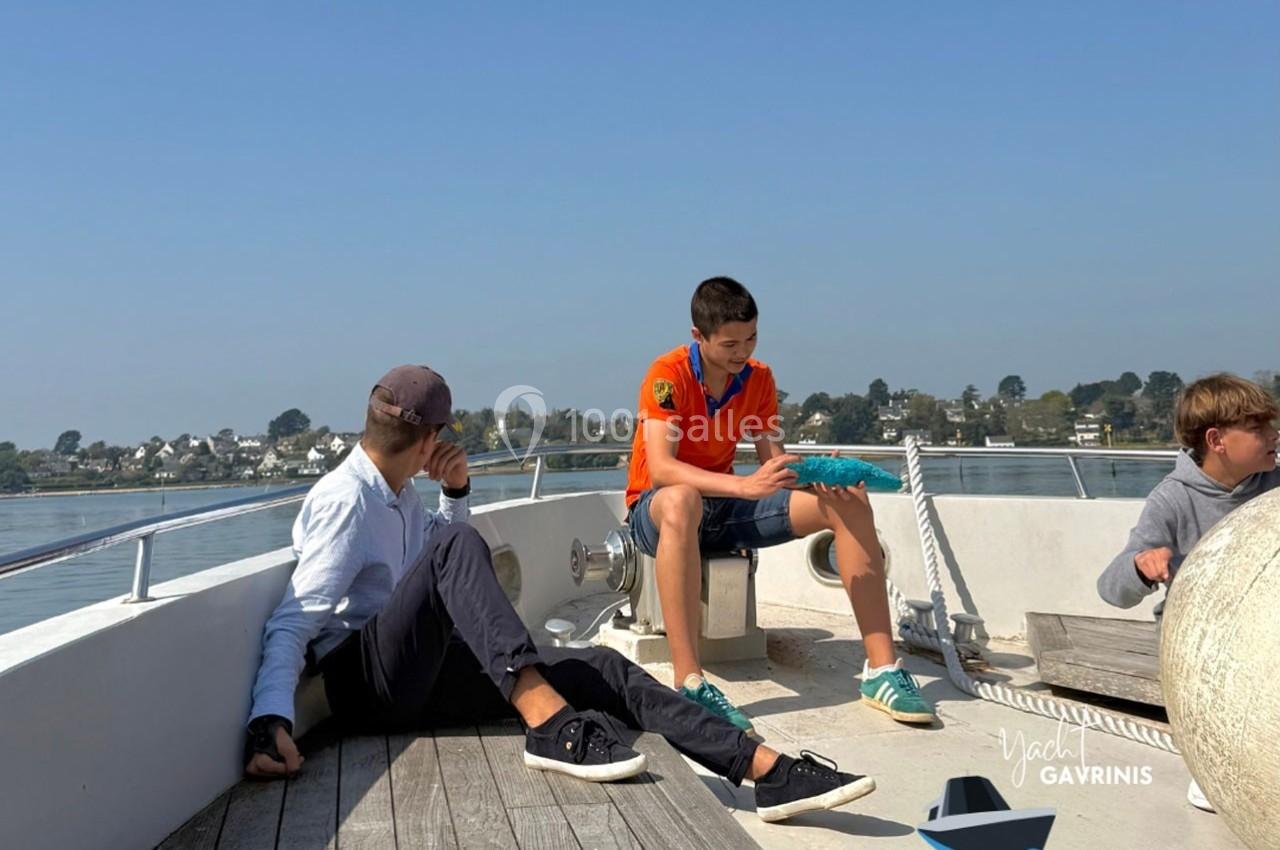 Deux adolescents et un homme assis sur le pont d'un bateau, avec un paysage côtier en arrière-plan.