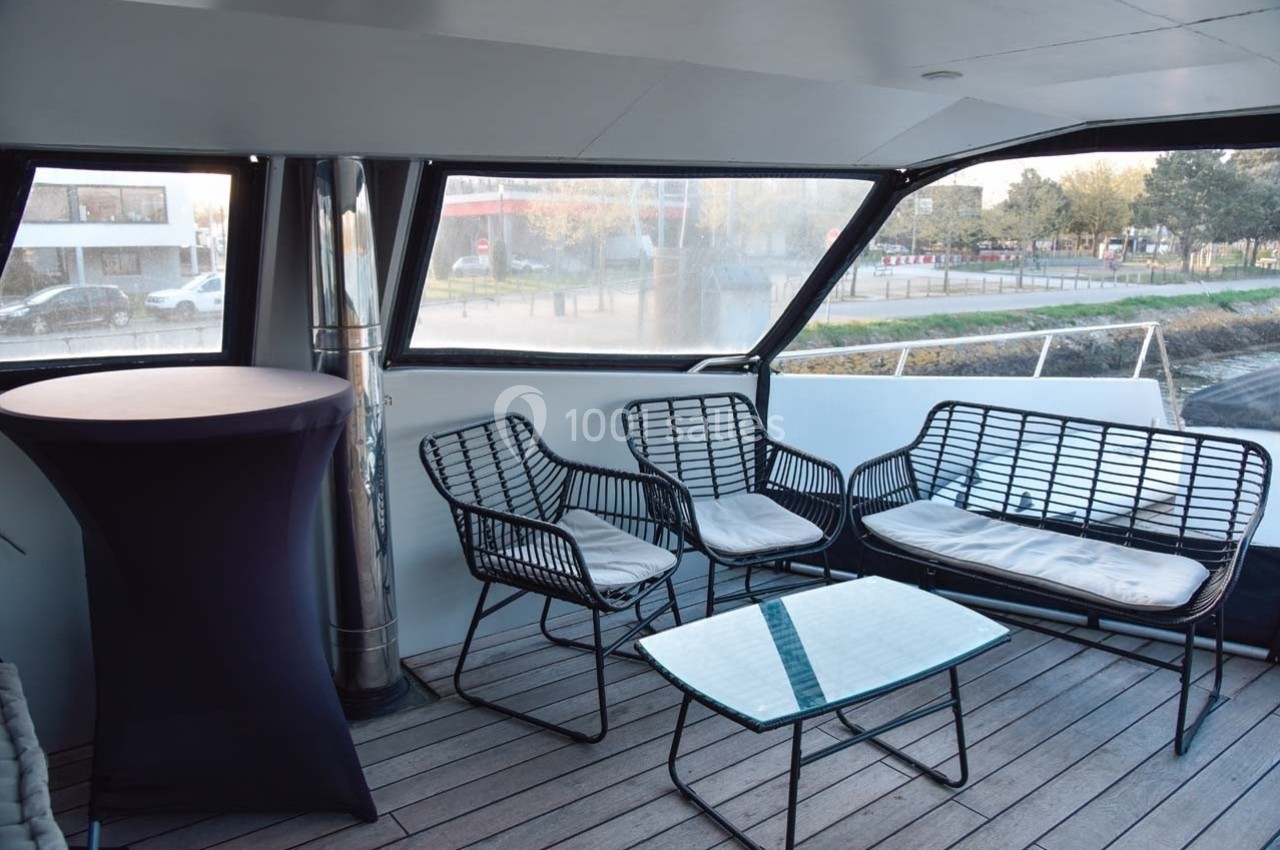 Espace extérieur avec mobilier en rotin noir, table basse en verre et vue sur un quai urbain.