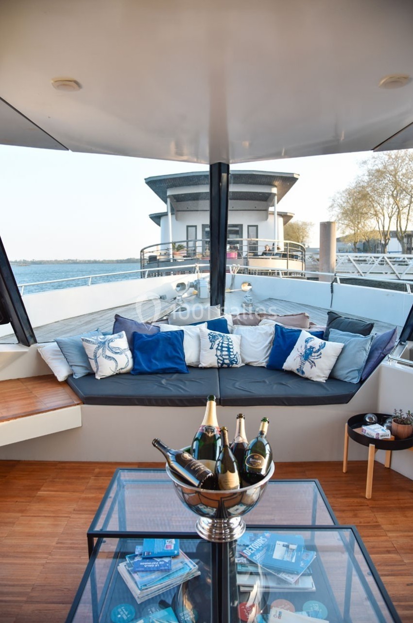Espace extérieur d'un bateau avec banquette garnie de coussins, table basse et vue sur l'eau et un bâtiment moderne.