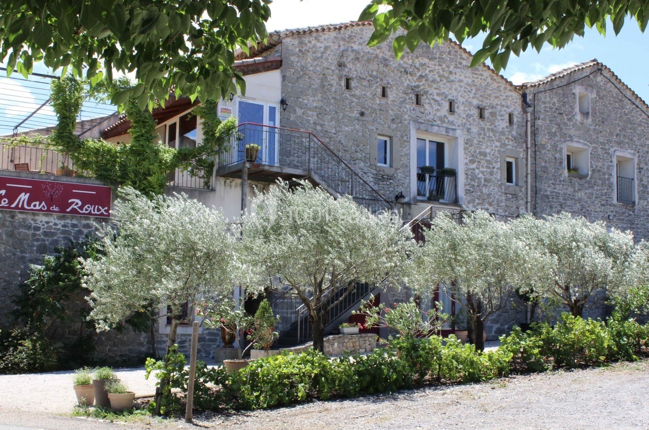 Location salle Quissac (Gard) - Le Mas de Roux #1 Façade d'une bâtisse en pierre entourée d'arbres et de végétation, avec un panneau indiquant ’Le Mas de Roux’.